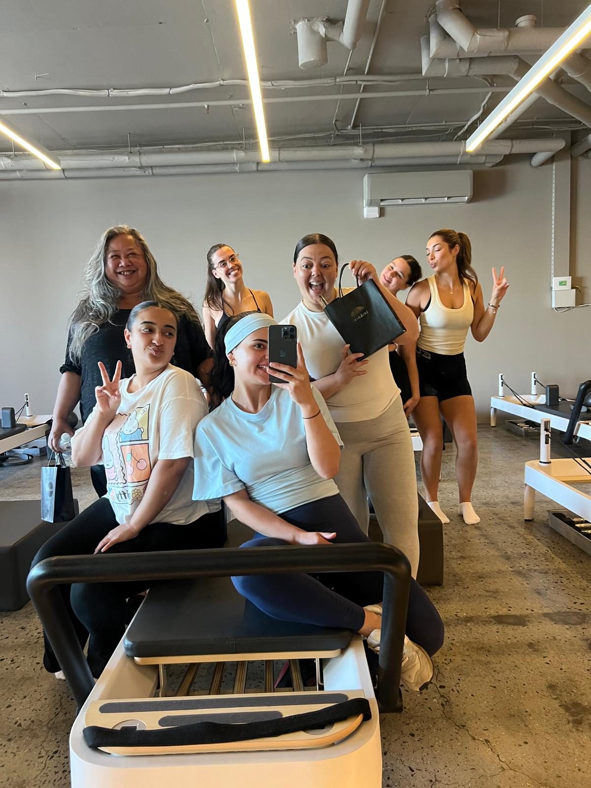 Lara takes a mirror selfie on a reformer with the AMS team gathered around her, pulling faces and holding up peace signs in the studio.