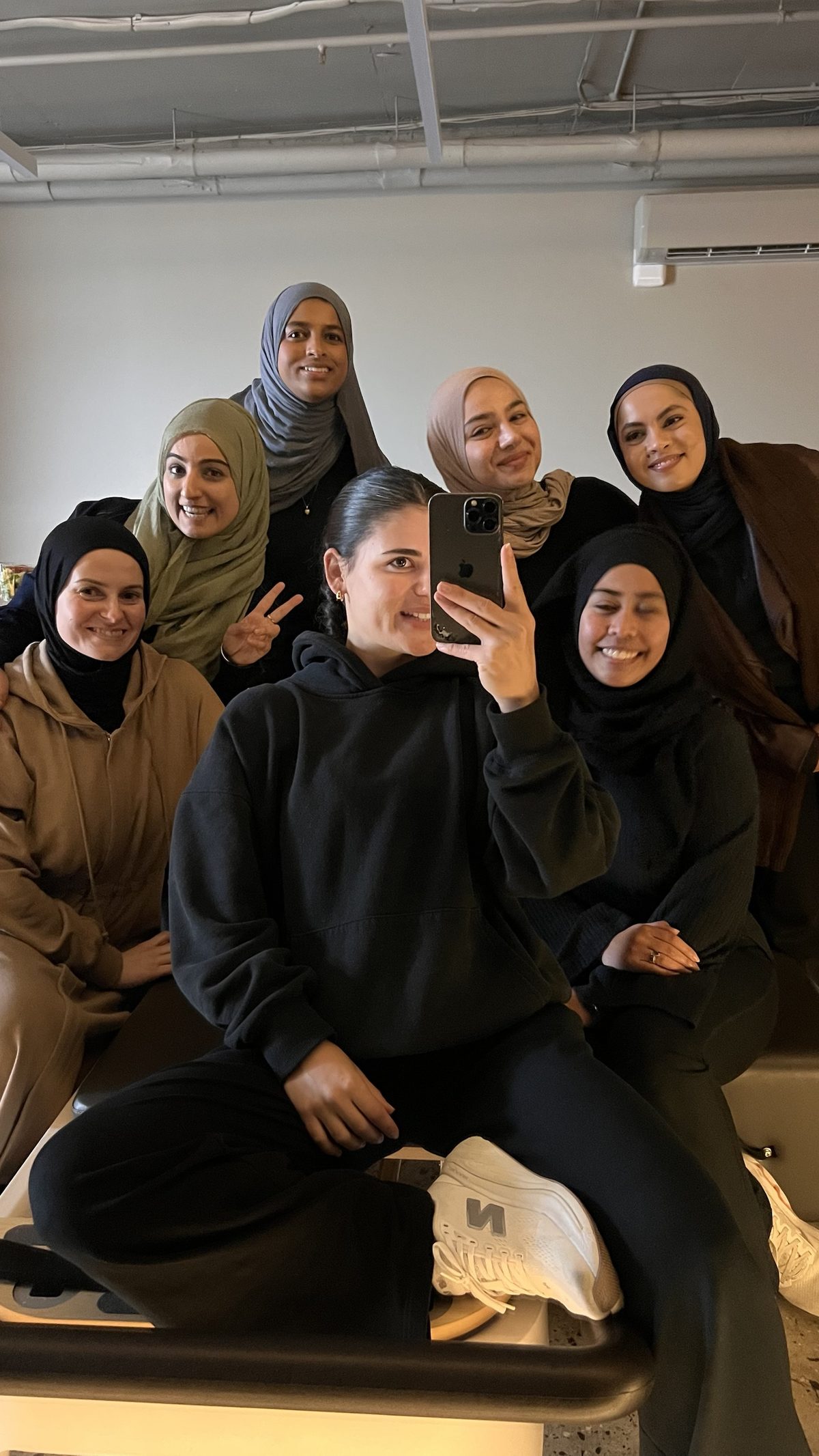 Lara, in a black hoodie, takes a mirror selfie while seated on a reformer, with six smiling women from Sisters Society gathered close around her in the studio.